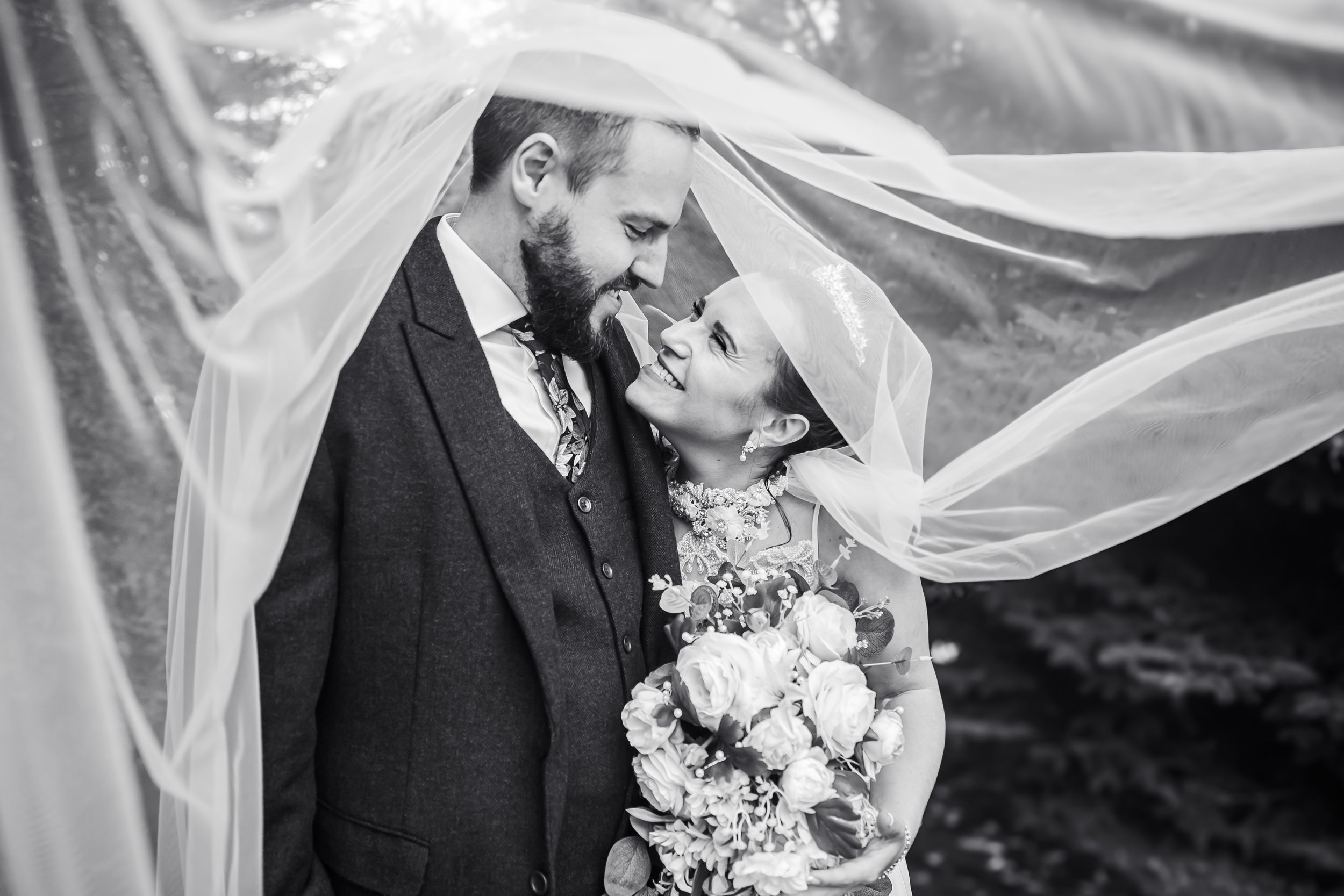 Black and white wedding photo of bride and groom smiling under flowing veil, bride holding bouquet.