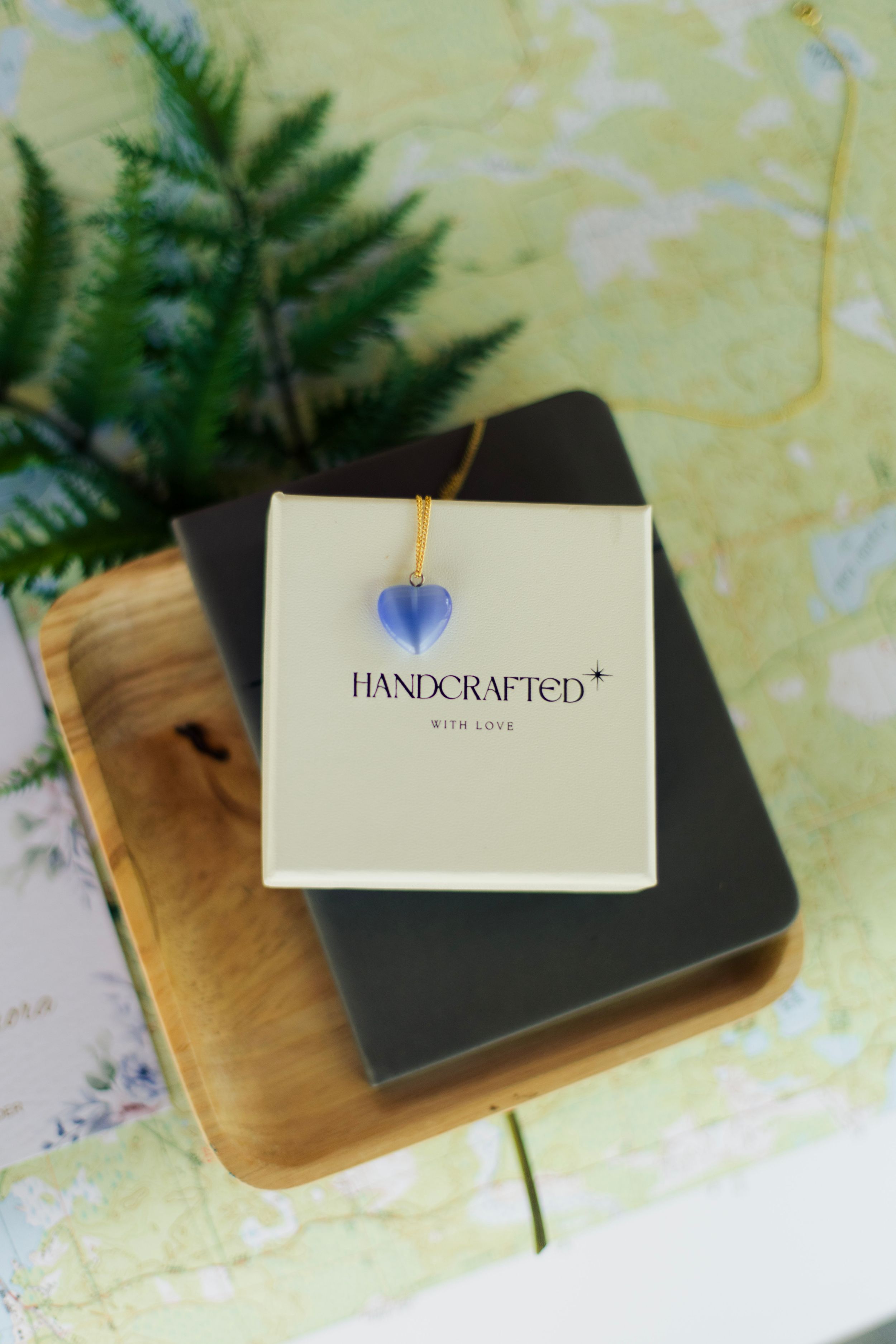 Blue heart pendant necklace on handcrafted jewelry box, displayed on wooden tray with fern and map background.