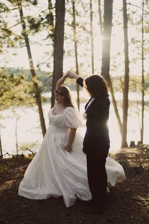 Bride in white gown and groom in black tuxedo dancing in sunlit forest near a lake.
