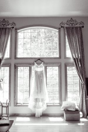 A wedding dress hanging on a hanger in front of a large arched window in black and white.