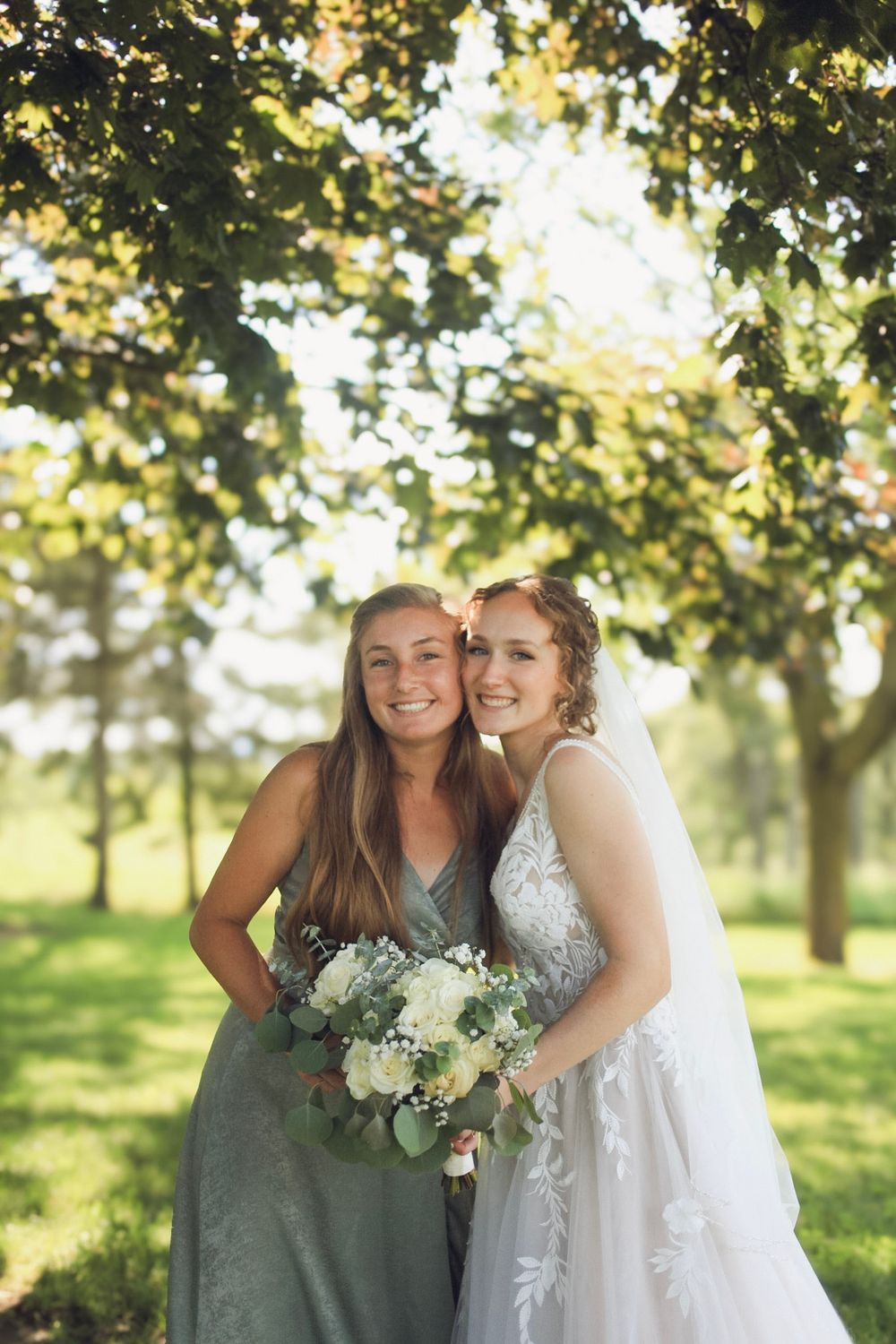 Bride in lace gown and bridesmaid in sage green dress holding white bouquets, posing outdoors under sunlit trees.