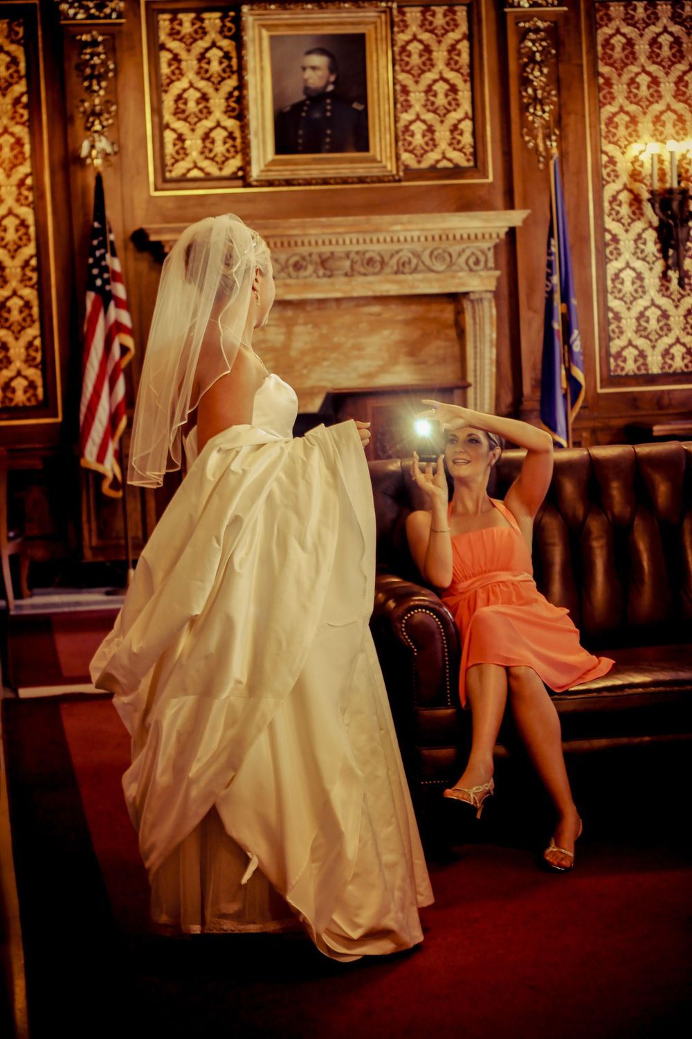 Bride in white gown poses while bridesmaid in coral dress photographs her in elegant room with fireplace and American flag.