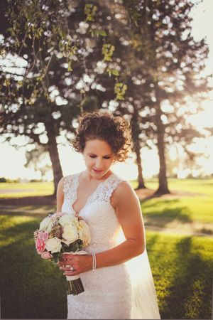 Bride in lace wedding gown holding pink and white bouquet, standing under trees in golden sunlight.