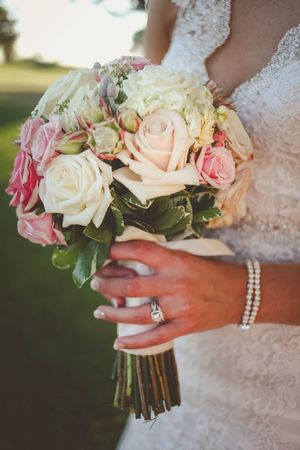 Bride holding a bridal bouquet of pink and white roses with pearl bracelet and diamond engagement ring.