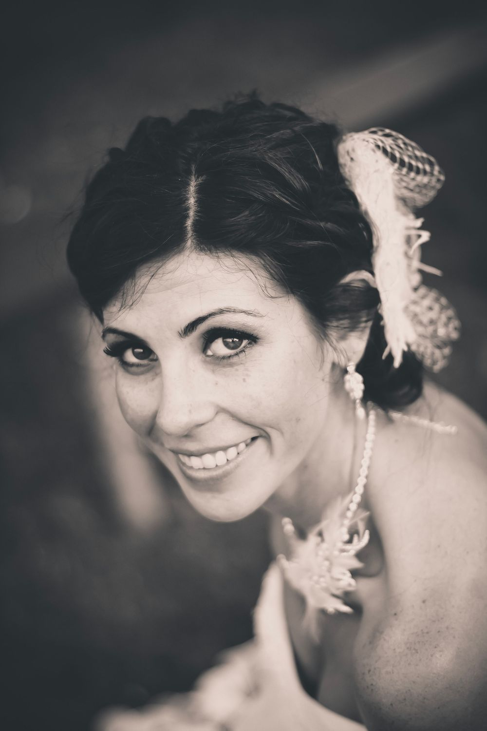 Black and white portrait of a smiling bride with dark hair, feather hair accessory, and pearl necklace.