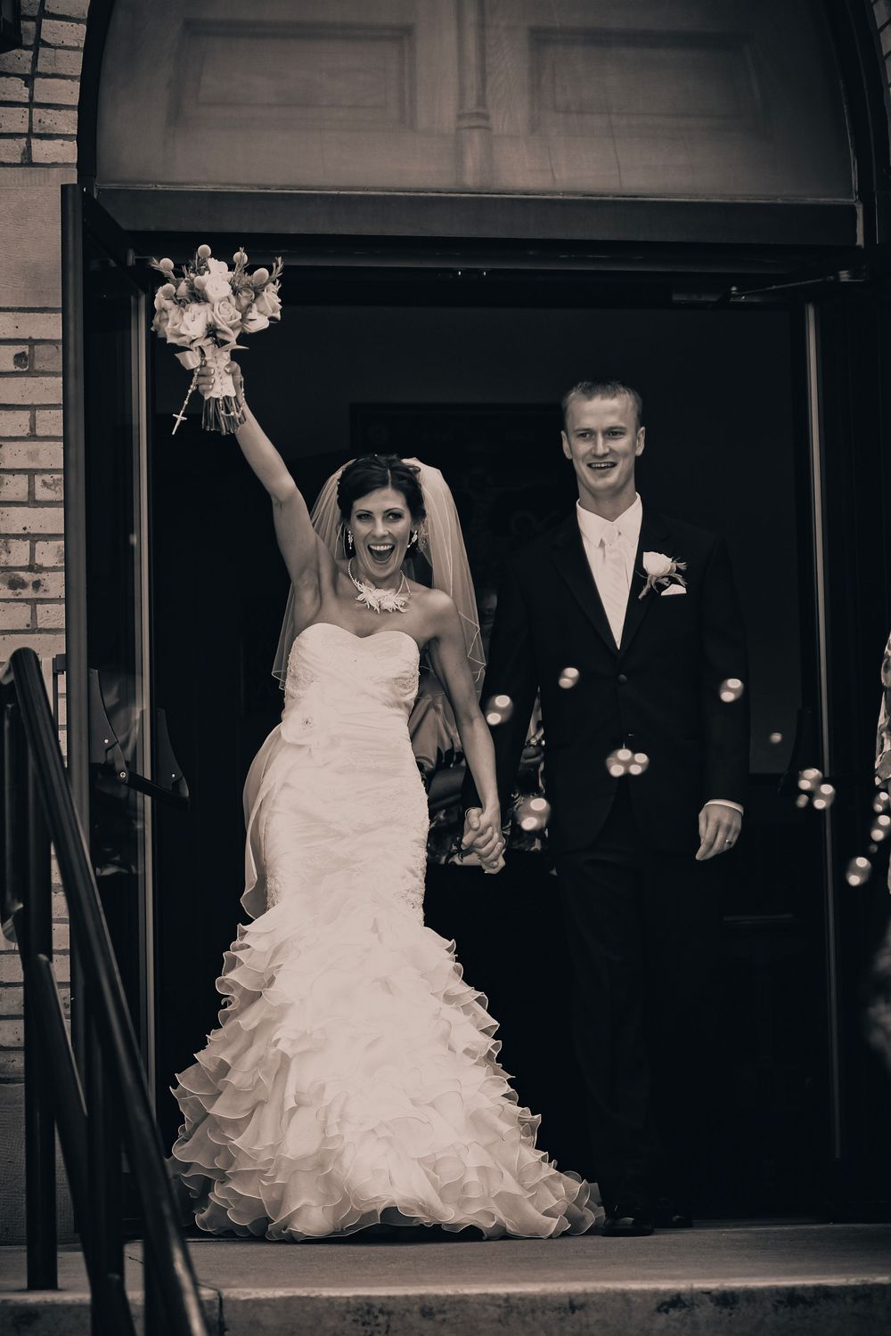 Black and white photo of a joyful bride raising bouquet and groom exiting church doors after wedding ceremony.