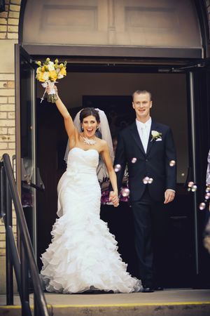 Joyful bride raising bouquet exiting church with groom after wedding ceremony, bubbles floating around happy newlyweds.