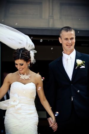 Smiling bride in strapless lace gown and groom in black suit hold hands as bubbles float around them after wedding ceremony.