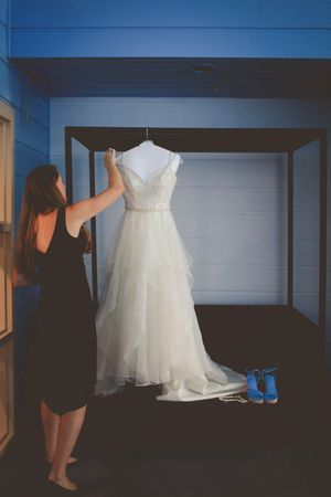 Woman in black dress admiring a white tiered wedding gown on a hanger, with blue shoes below.