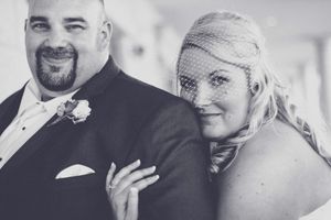 Black and white wedding photo of smiling bride in birdcage veil and groom in suit with boutonniere.