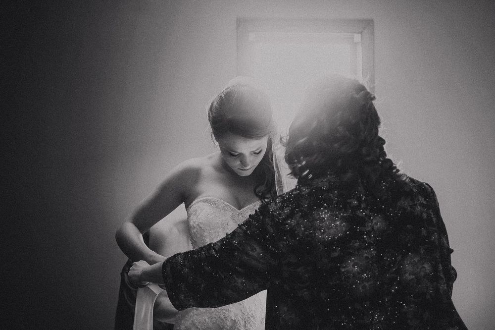 Black and white photo of a bride in a white gown being helped by another person, backlit by a bright window.