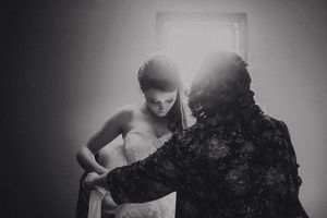 Black and white photo of a bride in a white gown being helped by another person, backlit by a bright window.