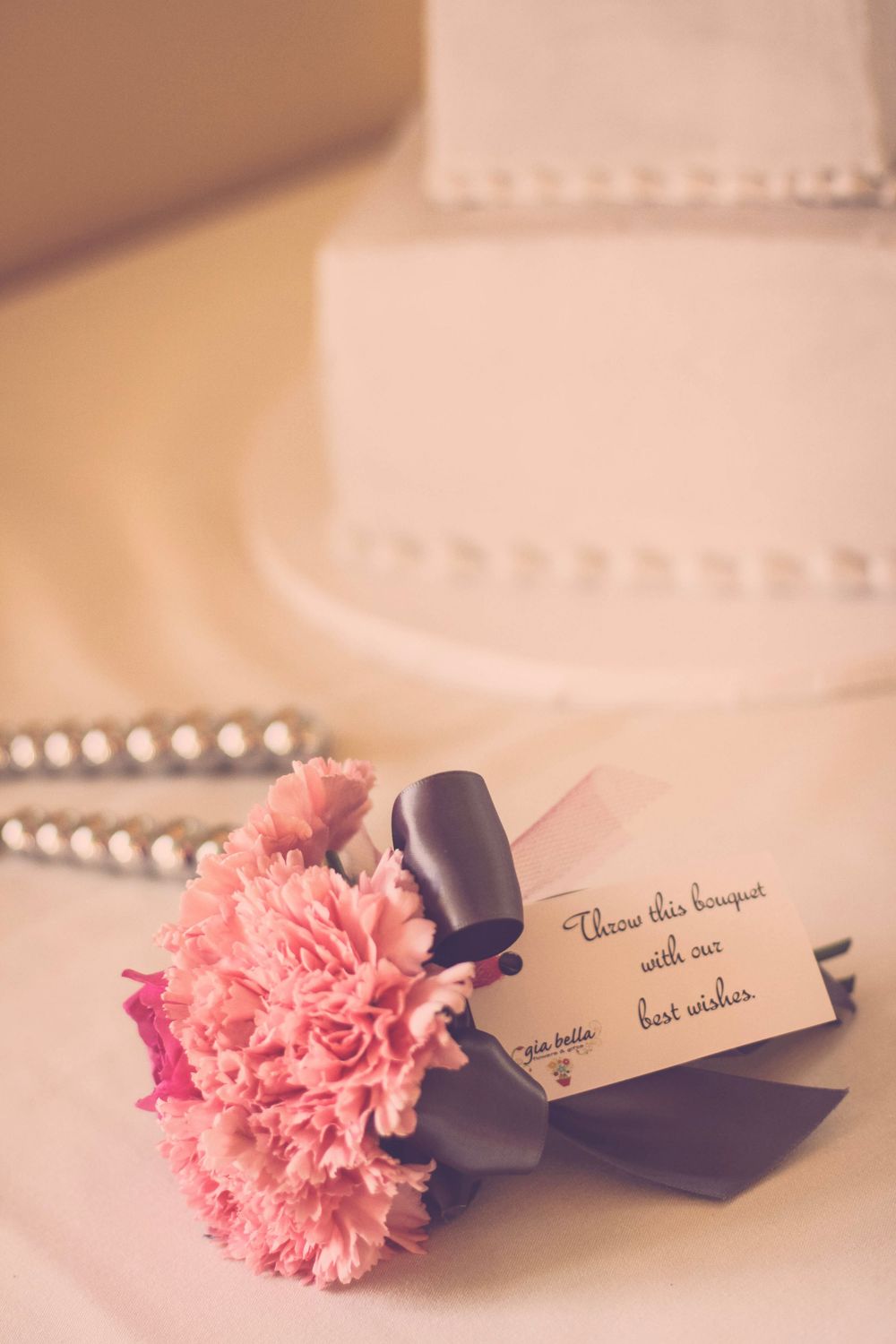 Pink carnation bouquet with tag reading best wishes beside a white pearl-decorated wedding cake.