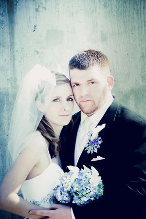 Bride and groom pose together on wedding day, bride holding blue floral bouquet wearing veil, groom in dark suit with boutonniere.