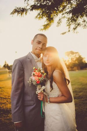 Bride and groom pose outdoors at golden hour, she holds a coral bouquet, both wearing wedding attire in a sunlit park.
