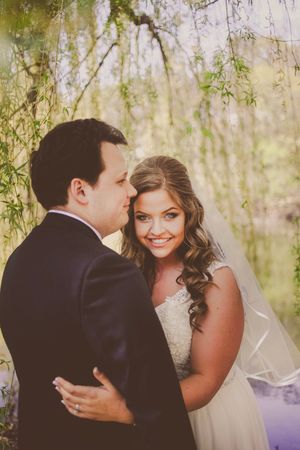 Bride and groom embrace under weeping willow tree, bride smiling at camera.