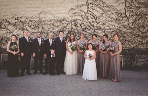 Wedding party posing outdoors in front of brick wall with bridesmaids in taupe gowns and groomsmen in dark suits.