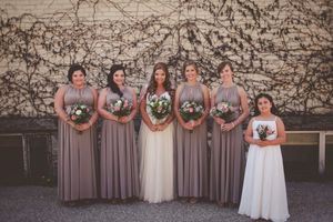 Bride in white gown with five bridesmaids in taupe dresses and flower girl holding bouquets posing outdoors.