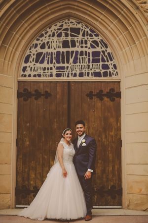 Bride and groom posing together in front of ornate wooden church doors with gothic arch and stained glass.
