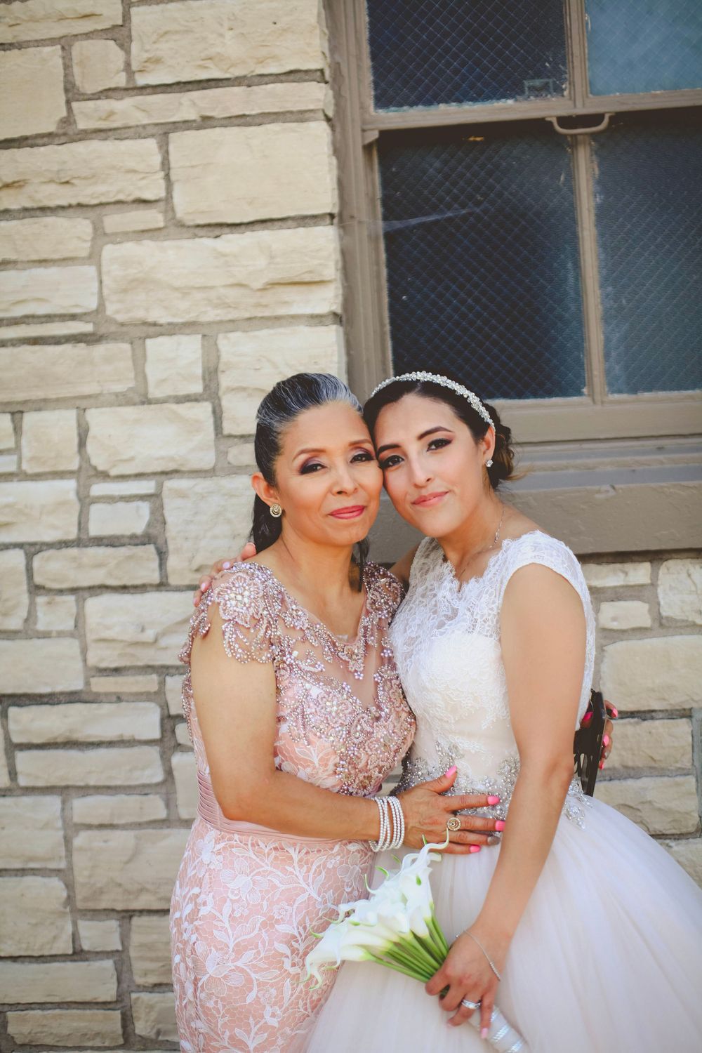 Bride in white gown holding calla lilies poses with older woman in pink lace dress against stone wall backdrop.