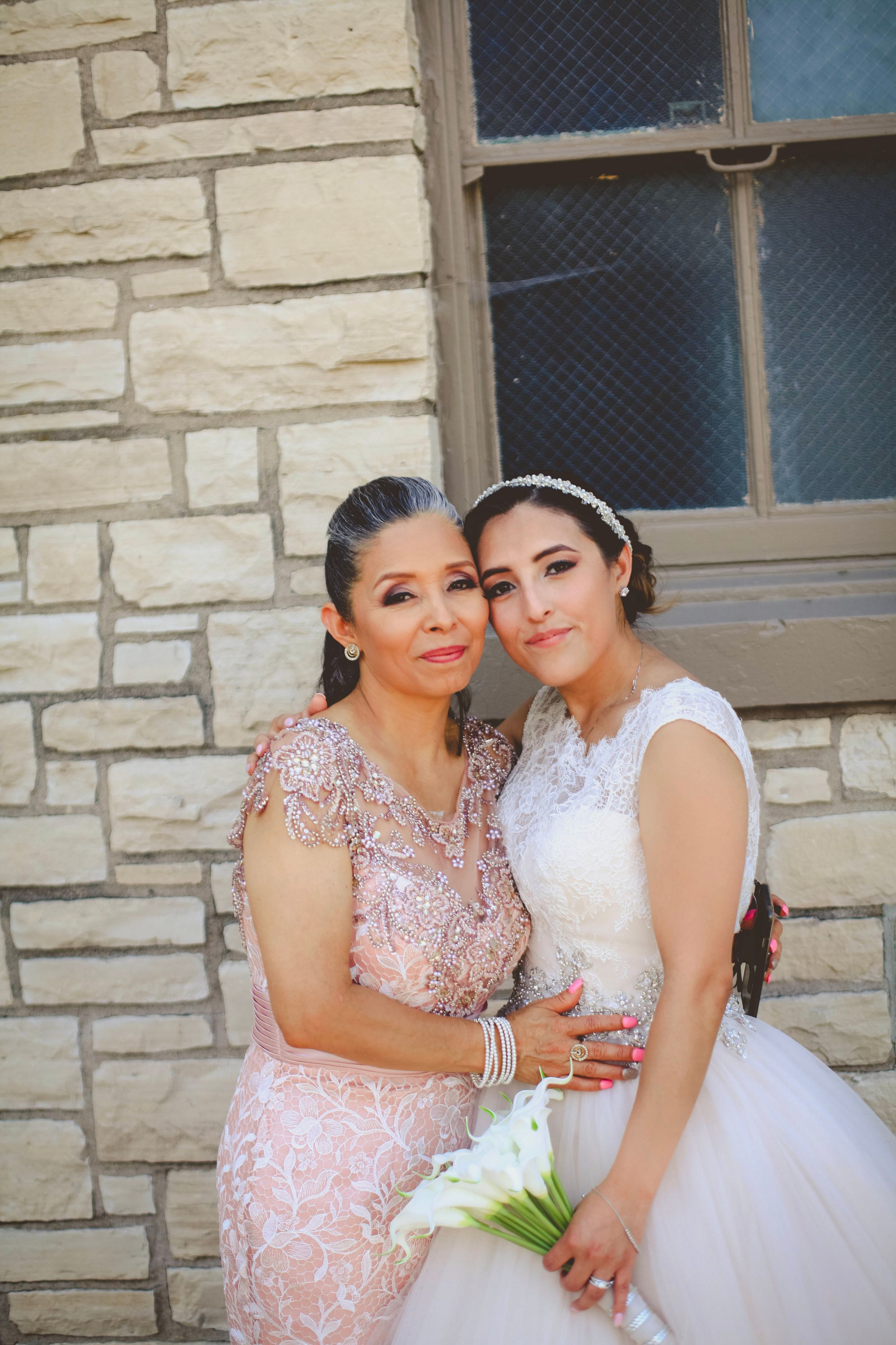 Bride in white gown holding calla lilies poses with older woman in pink lace dress against stone wall backdrop.