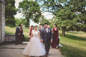 Bride in white ballgown and groom in gray suit walk hand-in-hand outdoors, with wedding party following on a tree-lined path.