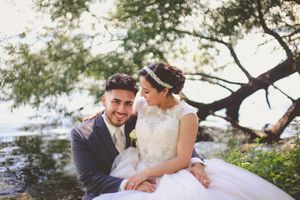 A smiling bride and groom sit together near a lakeside tree, the bride in a lace gown and groom in a gray suit.