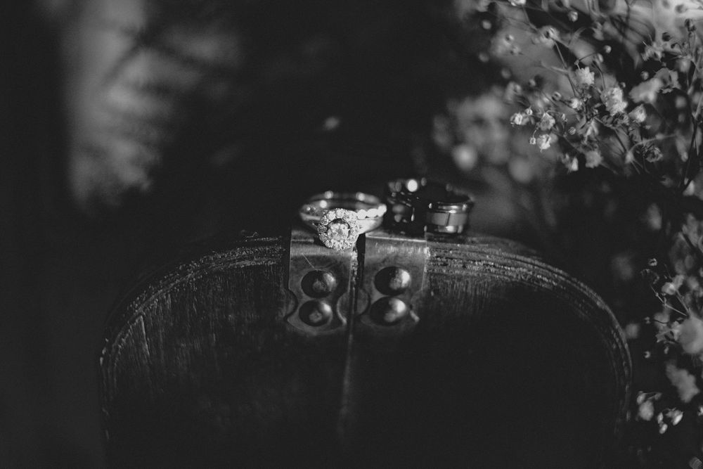 Black and white photo of vintage rings resting on an old leather camera with flowers in the background.