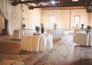 Elegant wedding reception hall with round tables draped in ivory linens, floral centerpieces, and rustic brick walls on hardwood floors.