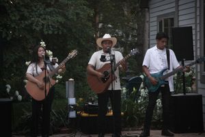 Three musicians perform outdoors, two playing acoustic guitars and one electric guitar, at a garden event.