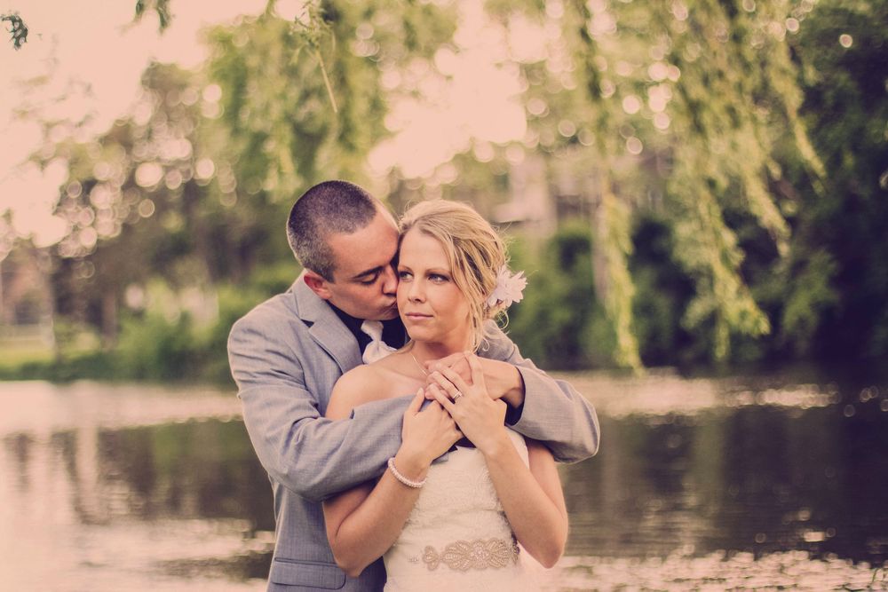 A groom in a gray suit embraces his bride in a white dress near a serene pond with lush green willow trees.