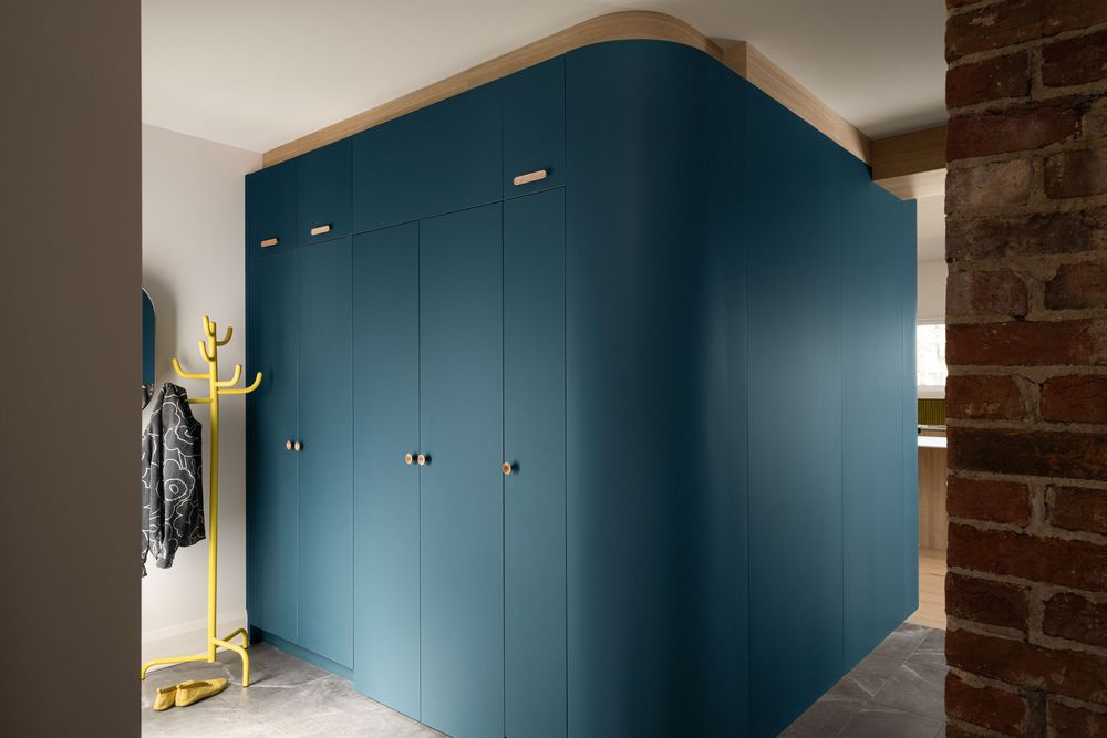Teal blue built-in wardrobe with brass handles in a modern hallway featuring exposed brick wall and yellow coat rack.