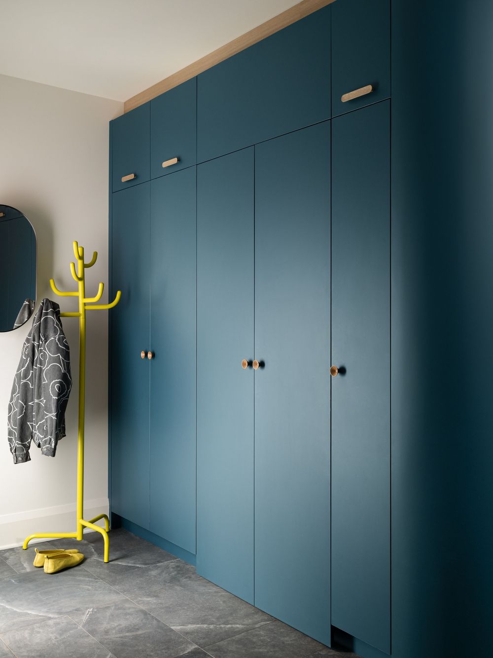 Teal blue floor-to-ceiling built-in wardrobe with brass handles next to a yellow coat stand in a modern entryway.
