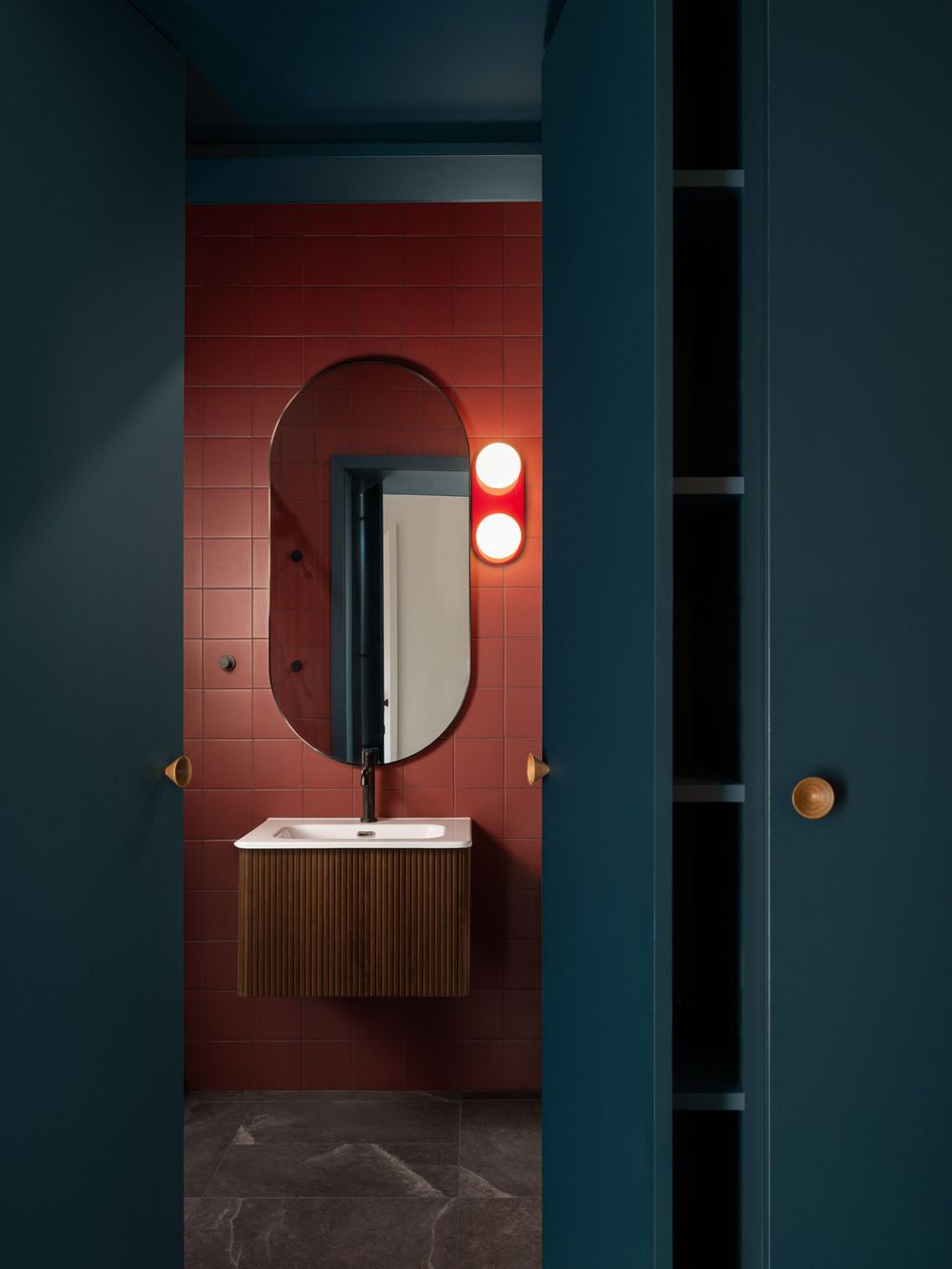 Modern bathroom with red tiles, oval mirror, globe wall sconces, and walnut floating vanity behind teal sliding door.