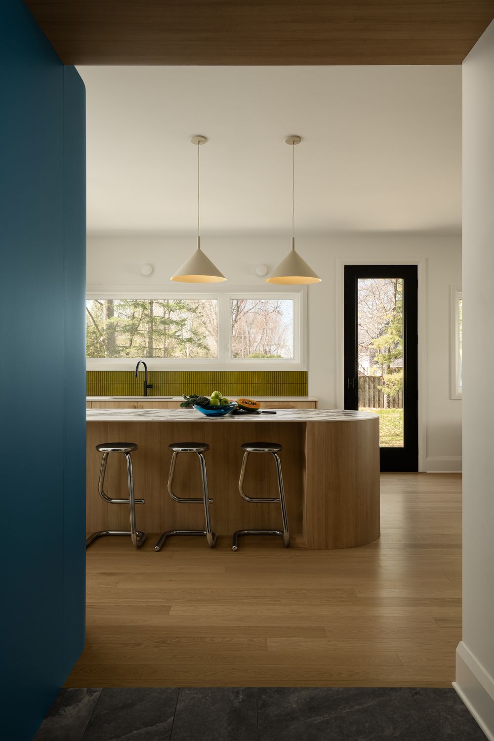 Modern kitchen with wood island, olive green tile backsplash, pendant lights, and chrome bar stools on light hardwood floors.