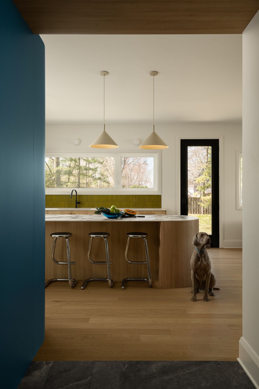 Modern kitchen with wood island, olive tile backsplash, pendant lights, chrome stools, and a dog sitting on hardwood floors.