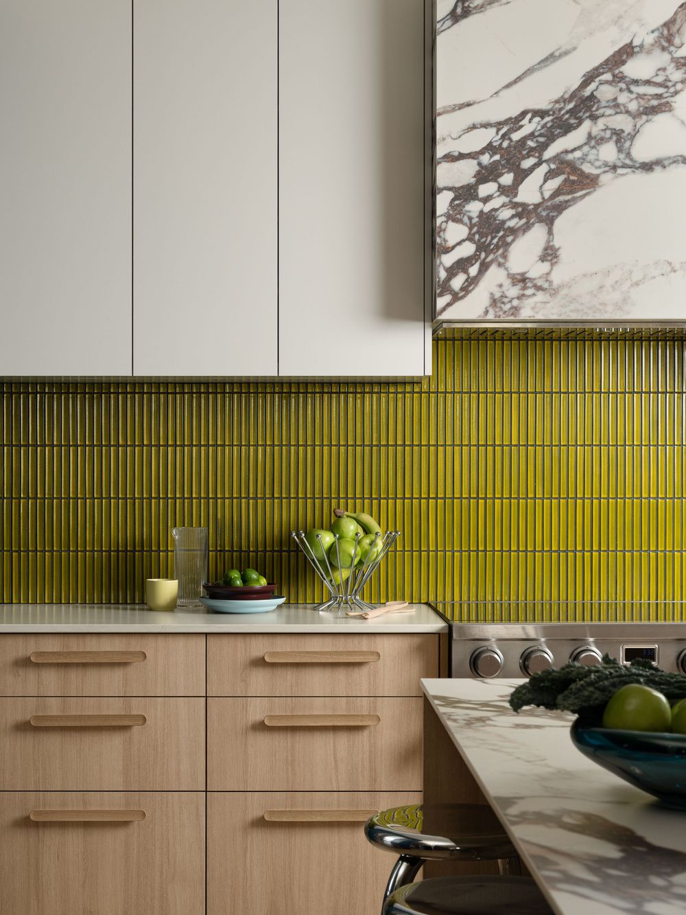 Stylish kitchen with chartreuse green vertical tile backsplash, light wood cabinets, white uppers, and marble hood surround.