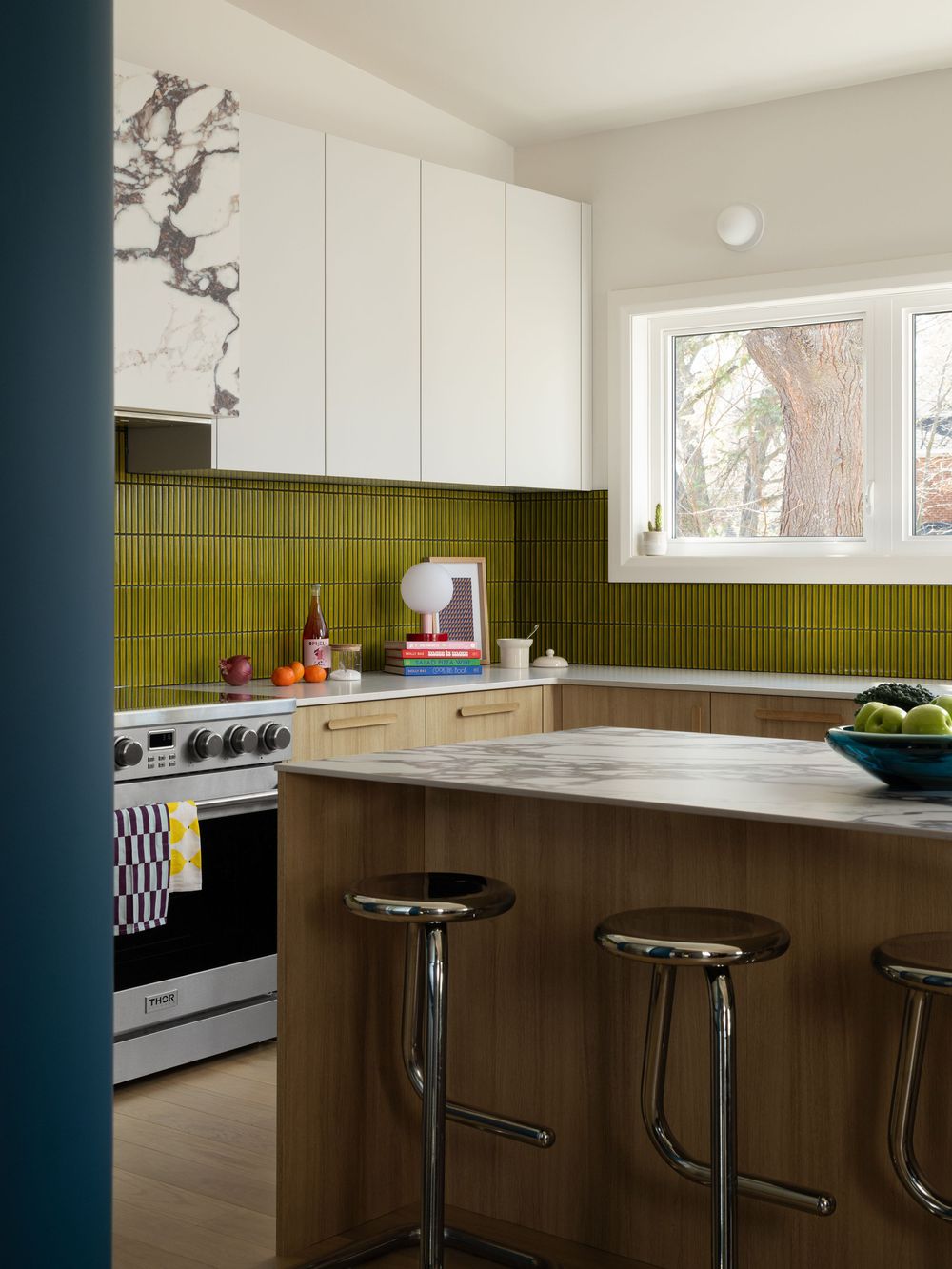 Modern kitchen with olive green tile backsplash, wood cabinets, marble countertops, and chrome bar stools.