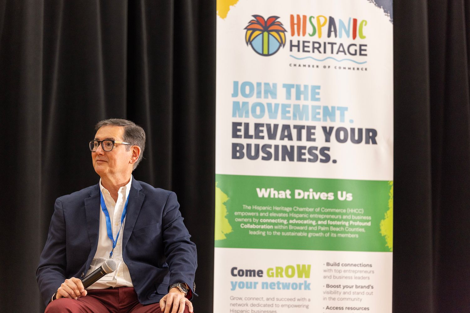 Man seated beside a Hispanic Heritage Chamber of Commerce banner reading Join the Movement, Elevate Your Business.