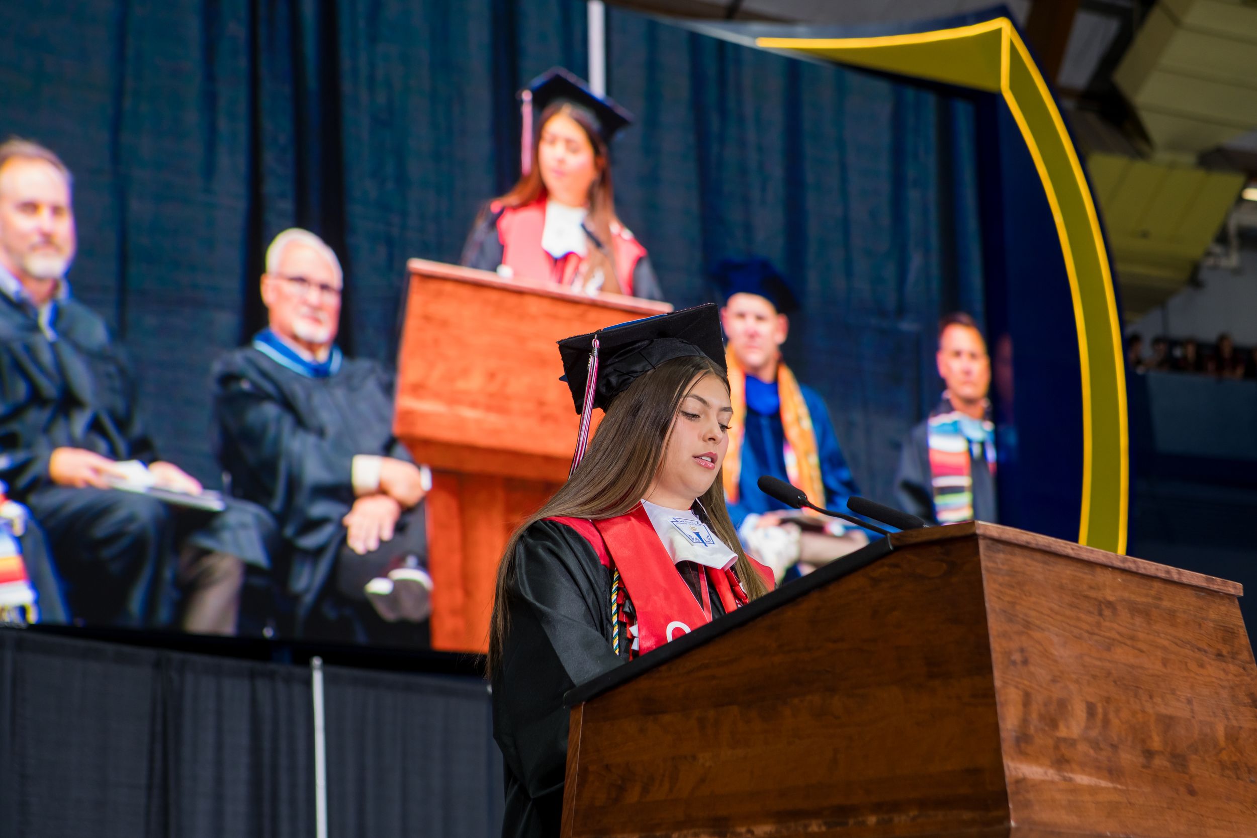 Nayeli Carbajal-Pettry in cap and gown speaks at podium during CHS commencement ceremony with faculty seated on stage.