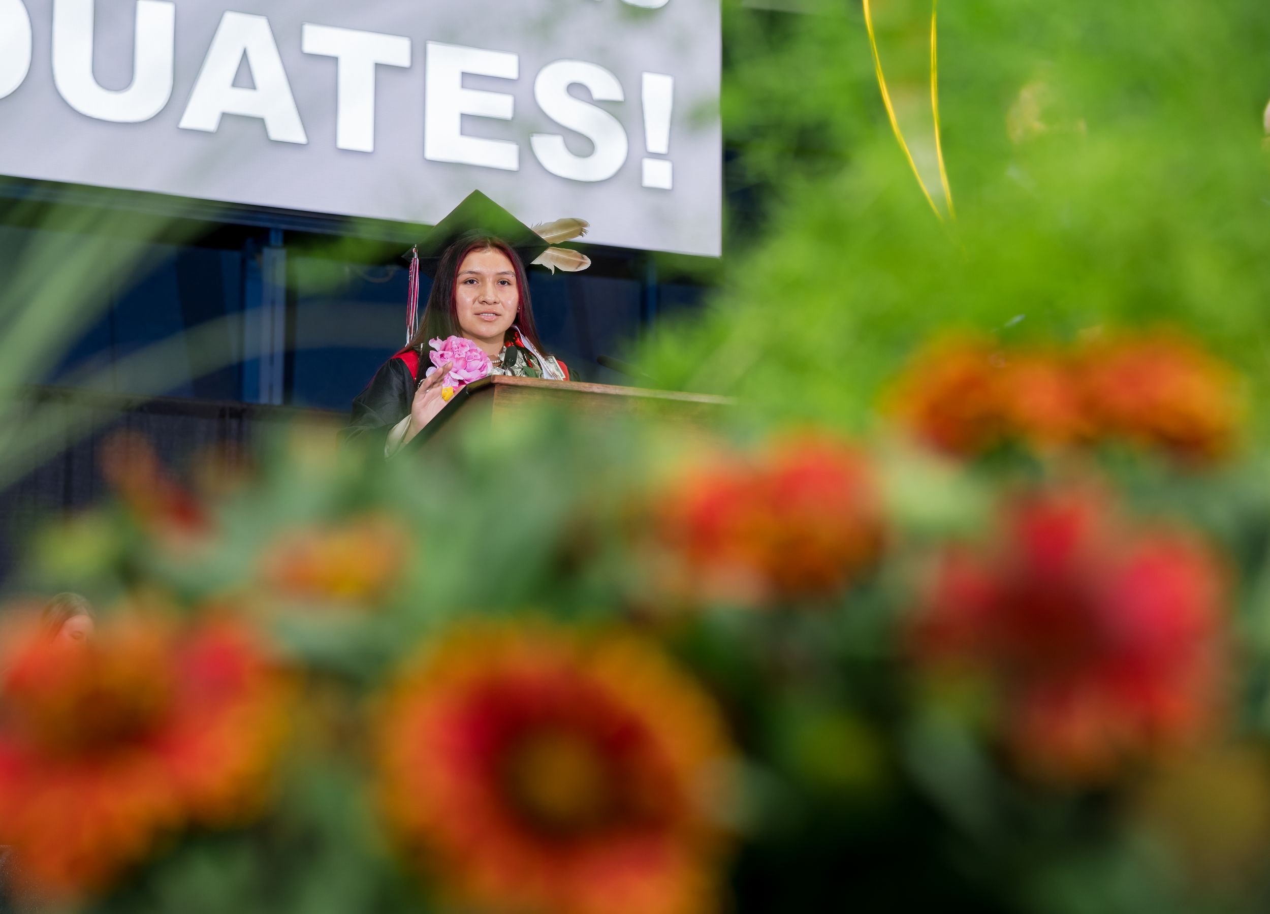 Graduation ceremony showing a speaker at podium with two graduates wearing red stoles on stage at commencement.