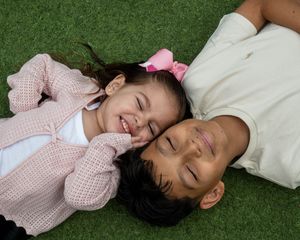 Two young children lying on green grass, smiling and laughing together, one wearing a pink cardigan and bow.