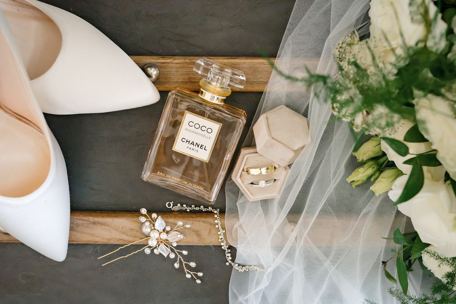 Chanel Coco perfume bottle with white bridal heels, wedding rings, floral hair piece, and white rose bouquet on gray surface.