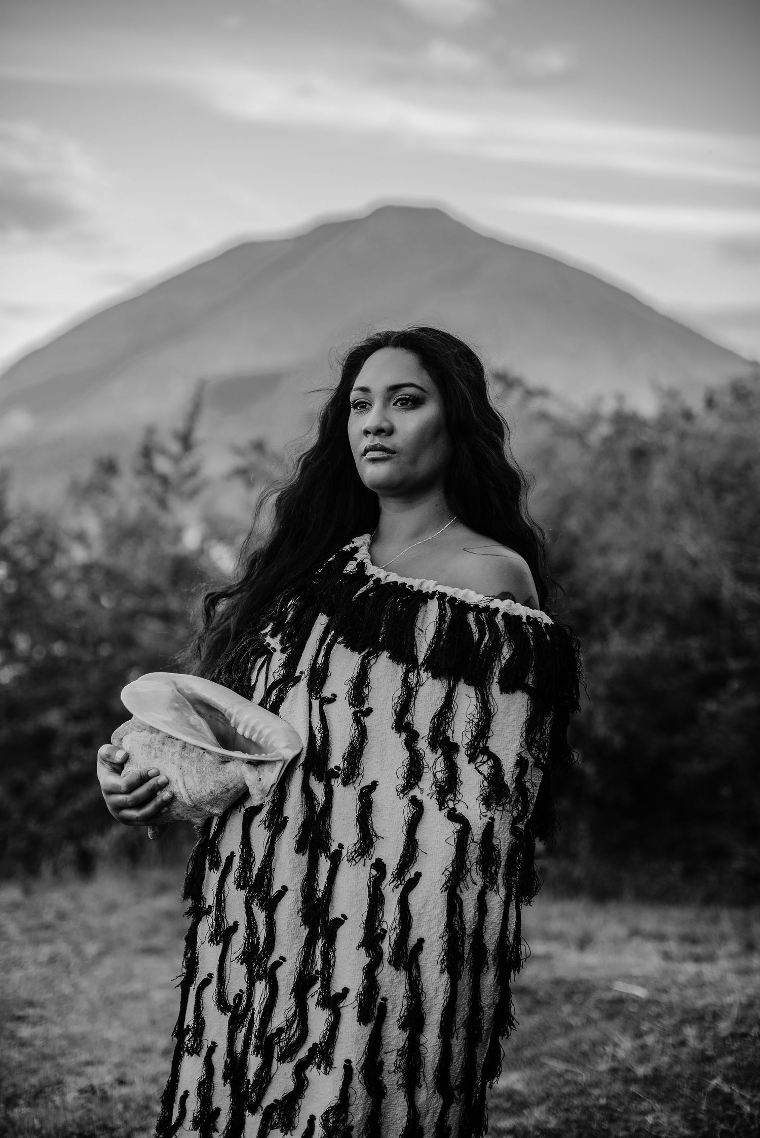 Person in traditional patterned dress stands before mountain landscape in black and white.