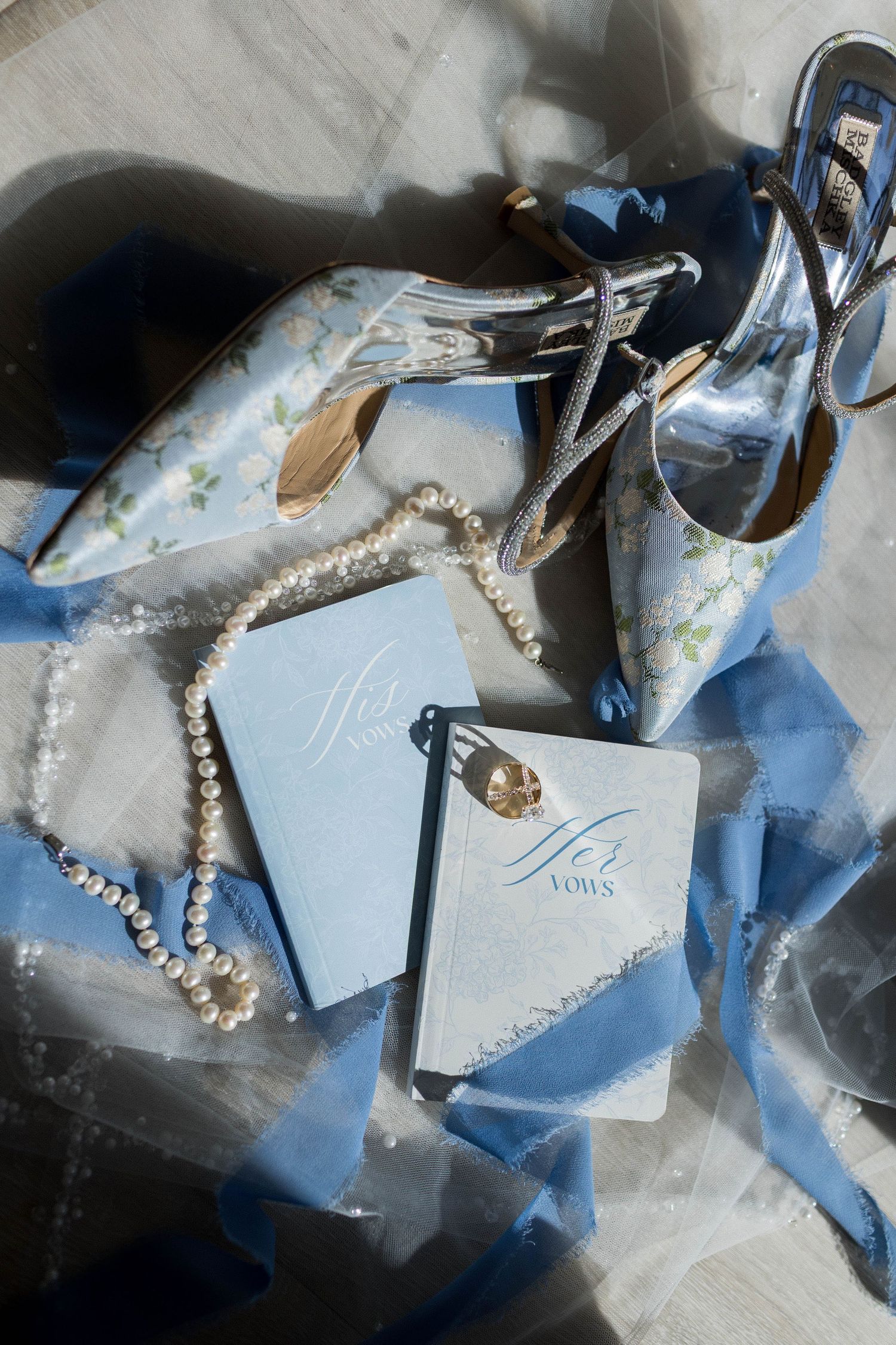 Blue wedding flat lay with floral heels, pearl necklace, vow books, and silk ribbon on a light surface.