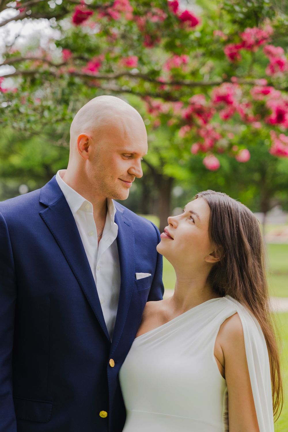 Couple in love gazing at each other outdoors, man in navy suit, woman in white dress, pink flowering tree backdrop.