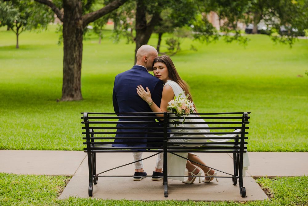 Couple in wedding attire sitting closely on a park bench, bride holding bouquet, surrounded by lush green grass and trees.