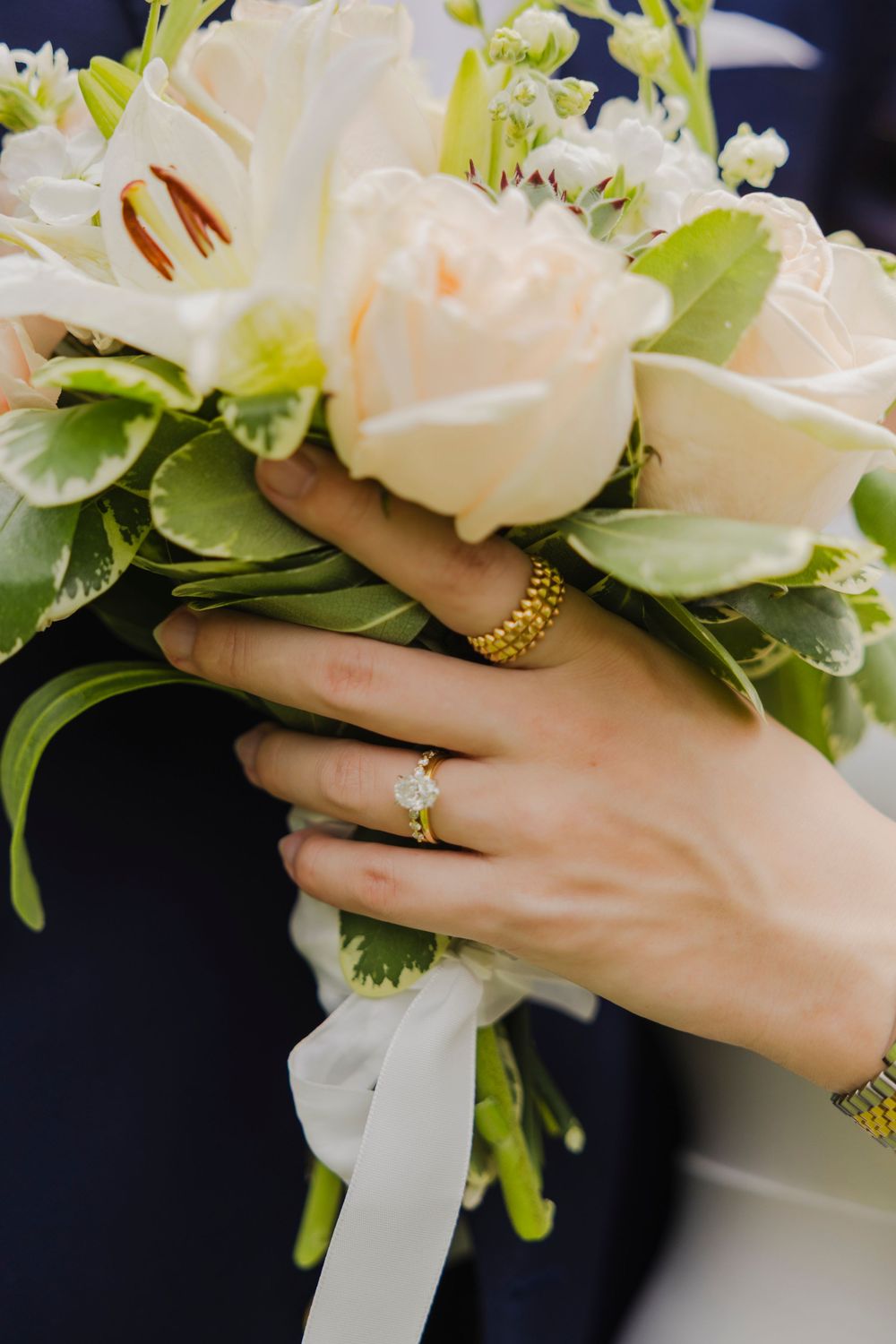 Bride holding a white rose wedding bouquet with green foliage, wearing a gold diamond engagement ring and bracelet.