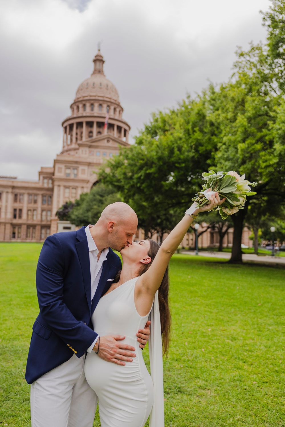 Couple kissing in front of Texas State Capitol building, bride in white gown holding bouquet aloft, groom in navy suit.