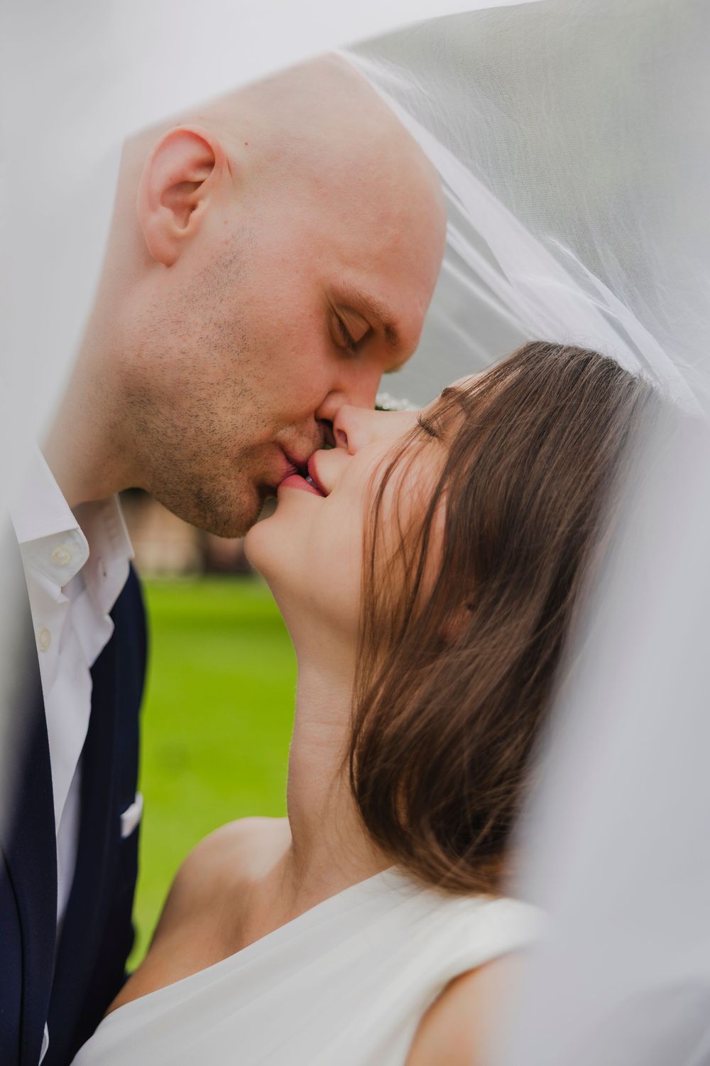 Bride and groom sharing a romantic kiss under a wedding veil outdoors on their wedding day.